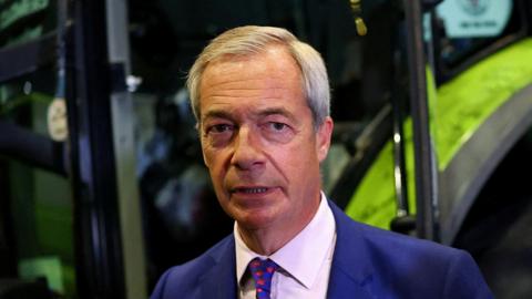 Nigel Farage wearing a serious expression standing in front of a mechanical digger