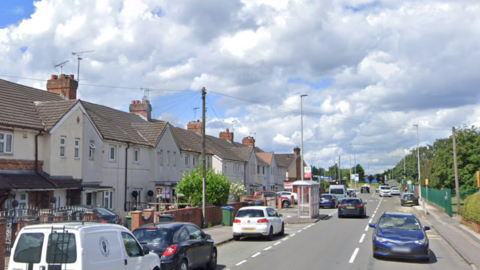 A street on a sunny day. There are white two-storey houses on the left side, and a bus stop on the pavement. There are trees on the right side. There are cars driving on both sides of the road.