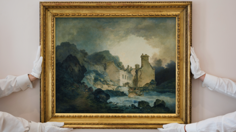Two pairs of hands holding up a framed oil painting by Turner. The painting shows a Georgian house amongst tree-lined cliffs. It is made of quite dark colours and has a moody sky.