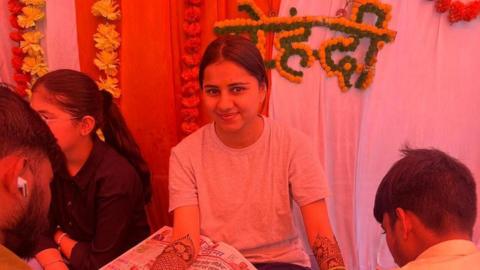 Harshita Brella in a grey T-shirt with henna decorations on her arms looks up at the camera and smiles without teeth in a room illuminated by a red light. There are two men drawing on her arms. 