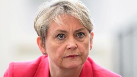 Home Secretary Yvette Cooper, a middle aged woman with short grey hair, stares intently at the camera. She is wearing a pink jacket