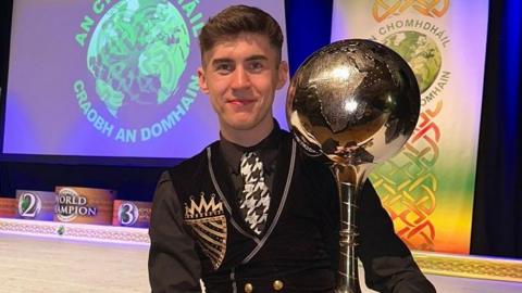 Barry Teague standing with his world championship trophy, a gold metal globe on a pedestal. Barry has short brown hair and is wearing a black long sleeved shirt with a black and gold waistcoat. He also has a white and black patterned tie on. It is an Irish dancing outfit.