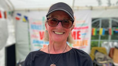 Stephanie Gray is wearing sunglasses and a black cap, smiling wearing a black top - standing in front of a marquee with colourful bunting and signage.