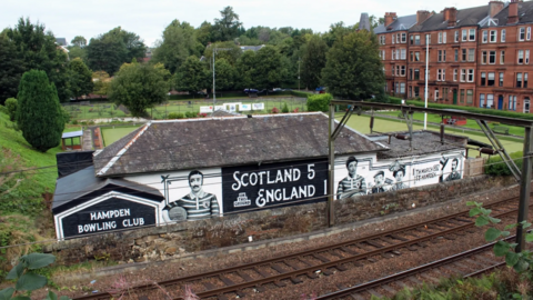 A bowling green ground from across train tracks behind it. A mural on the back wall says Scotland 5 England 1 with images of players from the 1880s alongside it. Flats can be seen in the background behind the green. 