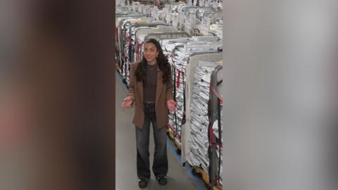 A woman in a brown coat and jeans stands next to a number of large trolleys containing exam papers.