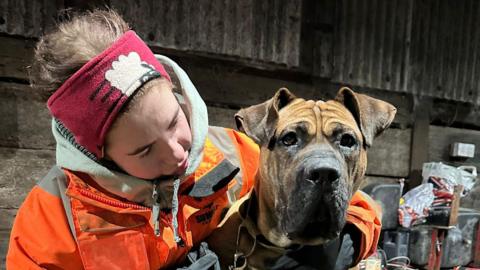 Michelle Halliday is wearing an orange hi-vis jacket and a red headband. She is looking at and hugging her cane corso dog Sparko, who has brown fur. They are inside a barn or similar building.