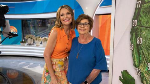 Savannah Guthrie in an orange top and patterned skirt, smiles and she wraps an arm around her mother, in a blue shirt and wearing glasses. They are on the Today set. 