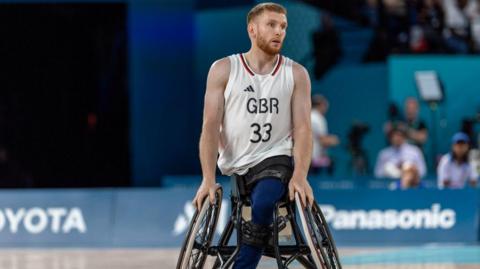 Ben Fox playing wheelchair basketball. He is wearing a white vest with GBR 33 on the front, and blue trousers
