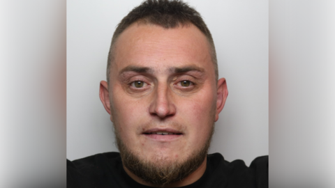 A mugshot of a man with short brown hair, a brown beared and a black top.