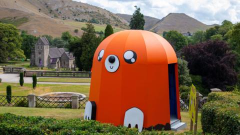 A large orange sculpture with a domed top is set within an open expanse of countryside - with hills and parkland in the distance behind.