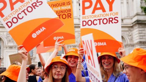Junior doctors protest in August 2023, wearing blue scrubs and orange hats hold placards reading: "Pay restoration for doctors".