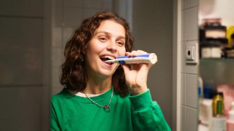 A woman smiles whilst brushing her teeth, looking at herself in the mirror.