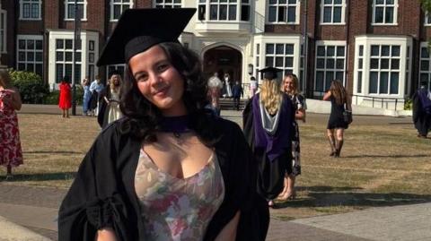 Mia Tanswell wears a graduation cap and robe while standing in front of a brown brick building and other university graduates