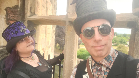 June Gothika is on the left she is wearing a black dress with some silver jewellery at her neck and a blue and black top hat while Gary Gimmick is wearing round steampunk black glasses and a black vest over a patterned shirt. They are standing against a stone window at Newark Castle.