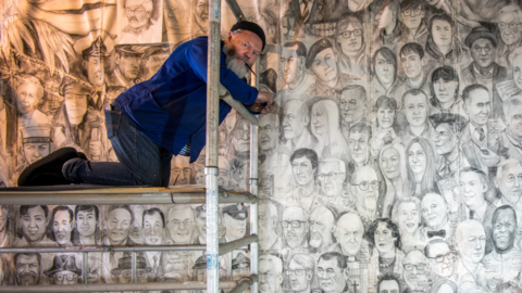 A man kneeling on scaffolding. He is sitting in front of a chalk mural. The artwork is made up of faces.