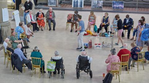 A large group of 20 or so older adults living with dementia, sat in a large circle on gold coloured chairs and two in wheelchairs. taking part in a musical session. Most participants are holding small percussion instruments.To the left of the images is a woman sat at a keyboard her mouth open in song or instruction, and has a large brass instrument next to her. A centre table hosts different musical instruments.