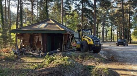 A wooden toilet block with a shallow pyramid shaped roof at the edge of a car park in a forest setting. Its sides are smashed and wrecked. A construction vehicle is next to it with smashed windows.