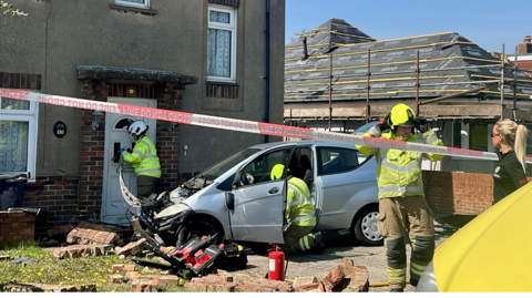 The image shows a car that has crashed into a house; its front is extremely damaged and firefighters can be seen securing the area. A paramedic in a black shirt is also on the scene.