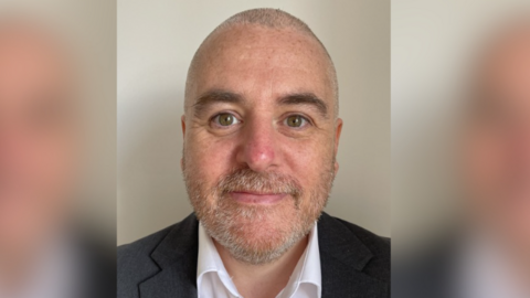 A close-up of Rob Heath, who is wearing a white shirt and black suit and has a moustache, stubble and hazel eyes.