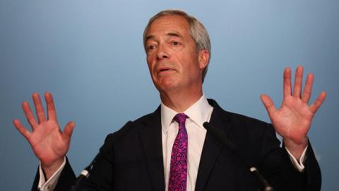 Nigel Farage looking slightly off camera. He has both of his hands raised at shoulder height with his palms facing towards the camera. He is wearing a black suit over a white shirt and a purple tie. He has greying hair and is standing in front of a blue background.
