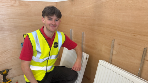 Flynn, who is a plumbing student at the HICSA, is wearing a red shirt and a yellow high visibility vest with the Sunderland College logo. He is smiling and has his left arm resting on a white radiator which is yet to be installed. 