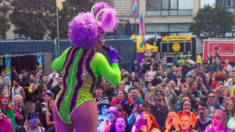 A singer wearing a green and purple leotard and a purple wig is sings to a crowd at Newcastle Pride. 