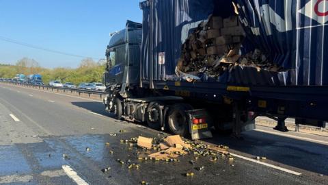 A lorry with the side of its trailer ripped. Boxes of wine are visible, with some smashed and crumpled in the road. The carriageway is clear of traffic, though there is a queue on the other side.