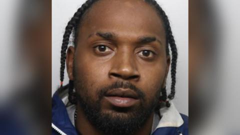 Mugshot of Ranei Wilks looking straight at the camera and wearing a blue and white jacket. He has black hair tied in braids and has a short dark beard. 