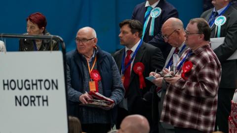 Labour party council candidates and supporters listen to declarations in the Sunderland Council local elections at Silksworth Community Pool. There are four men wearing red rosettes looking to the left, some of them are taking notes. There are two people in suits behind them wearing turquoise rosettes for Reform. 