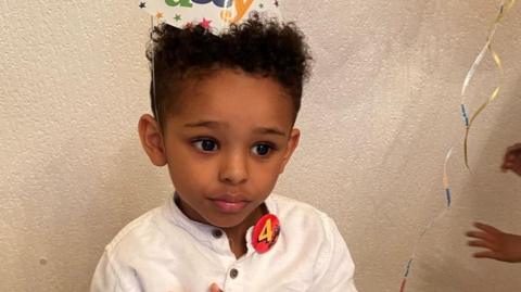 Fareed Amir on his fourth birthday, wearing a "4" birthday badge, a party hat and a white shirt.