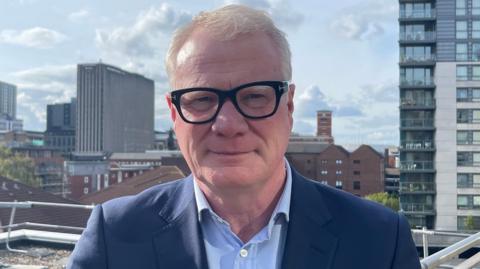A man looks into the camera with a serious face, and a slight smile. He has thick black glasses on with grey/white hair. He is wearing a blue blazer and a blue top. There are skyscrapers behind him. 