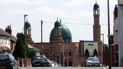 A street view of Makkah Masjid in Leeds. It has a large green dome and a striped brick design.