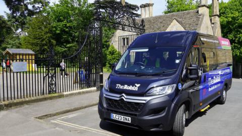 A  navy mini bus is parked near a black gate with a park in the background and cream-coloured homes.