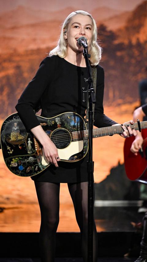 Phoebe Bridgers performs on stage holding a black, painted guitar. She is a young woman with short, blonde hair. She is wearing a black dress and she is singing on stage. 