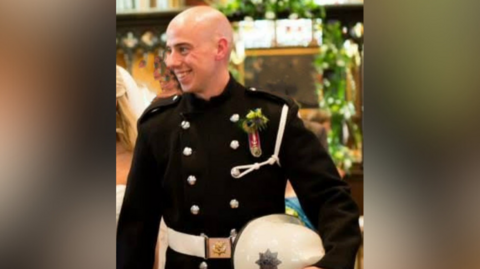 A picture of Martyn Sadler on his wedding day. He is wearing a black uniform and is smiling, looking away from the camera. A woman in a wedding gown could be seen walking behind him.