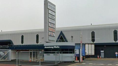 Exterior of the factory, which is large and grey, and the guardhouse which is grey with razor wire around the top. Next to the guardhouse is a tall tower with signs naming the site as Armstrong Works and other company names on it.