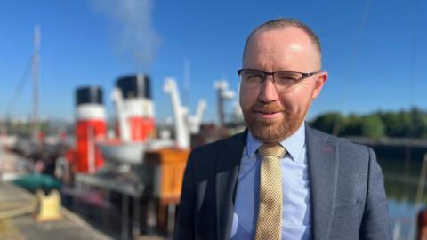 Paul Semple, General Manager of Waverley Excursions, smiles at the camera while standing in front of the paddle steamer. He wears a navy suit with a blue shirt and yellow tie. 