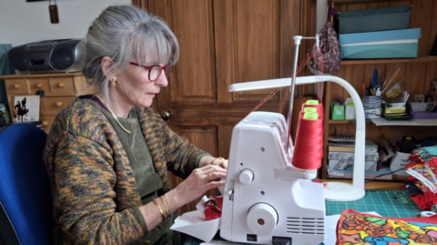 A woman with grey hair and a brown patterned cardigan sitting beside a white sewing machine with wooden cupboards behind her