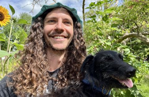 A man with long curly dark hair holds a small black dog. The dog has its tongue out and the man is smiling.