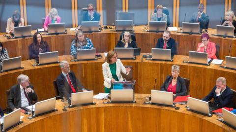A group of Senedd members sat in the Senedd debating chamber, with the first minister Eluned Morgan stood at the front in the centre, in a white jacket and green dress.