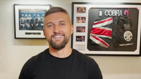 Man in black t shirt smiling. He's standing in front of two pictures showing boxing memorabilia
