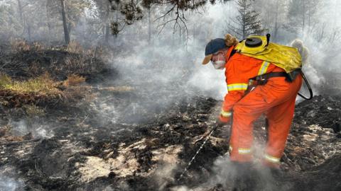 A person wearing orange, protective overalls and wearing a face mask and cap uses water to dampen down hotspots at RSPB Scotland's Loch Garten reserve at Abernethy.