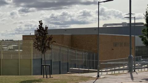 An outside view of HMP Five Wells prison. It shows a large brick building surrounded by metal fencing. 