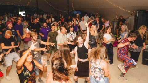 A group of people dancing underneath a marquee. It is dark outside and there are strings of lights draped across the top of the marquee.