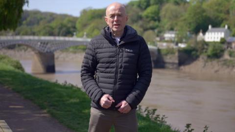 Gareth Lewis standing on a riverbank with a bridge in the background