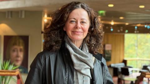 A woman with curly brown hair smiles at the camera. She is wearing a black leather jacket and grey scarf. There is a cafe behind her. There are large photos on the wall.