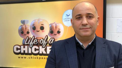 Mahmut Gunaydin wearing a dark suit.  He is standing in front of a screen showing an image of his schools' programme "Life of a Chickpea".  The image shows three chickpea characters and corresponding text.