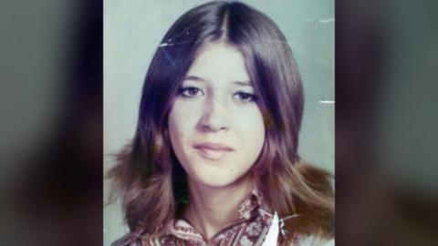 Laura Aimee, in a school photo from the 1970s. She has long brown hair parted down the middle and brown eyes, and wears a patterned blouse.
