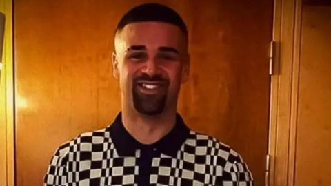 Tariq Evans smiles at the camera wearing a black and white checked polo shirt. His black hair is short and he has a short beard and moustache.