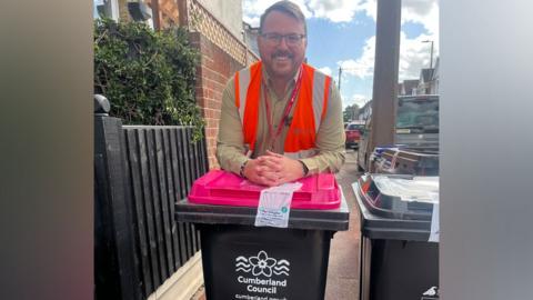 Daniel smiles at the camera while leaning on the bin, which is black with a pink lid. It has a white logo of a flower with waves either side and the words Cumberland Council. Daniel wears a high vis jacket over a beige shirt and has a red lanyard round his neck. He is standing on a pavement where there are other bins and also parked cars.